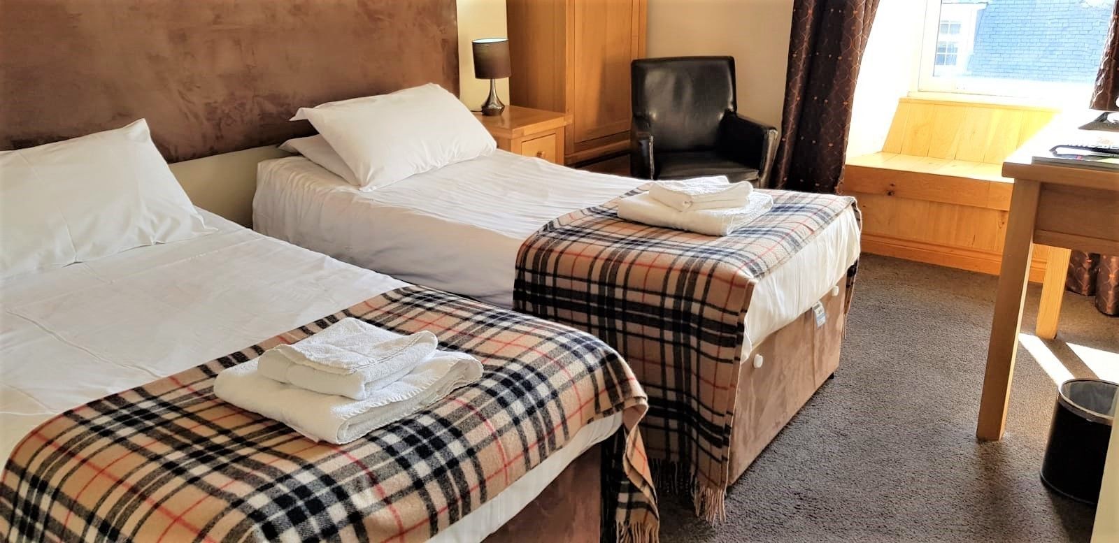 Two single beds with white linens and plaid blankets, a wooden nightstand with a lamp, a black chair, and a wooden bench by the window.