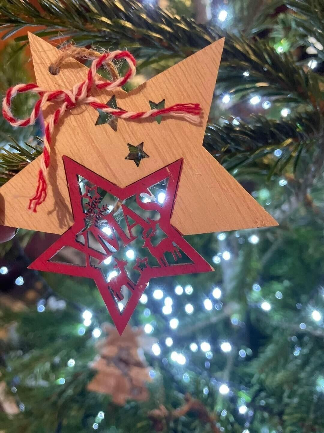 Un ornamento a forma di stella di legno con un'apertura a stella rossa, legato con un cordoncino rosso e bianco, appeso a un albero di Natale addobbato con luci.