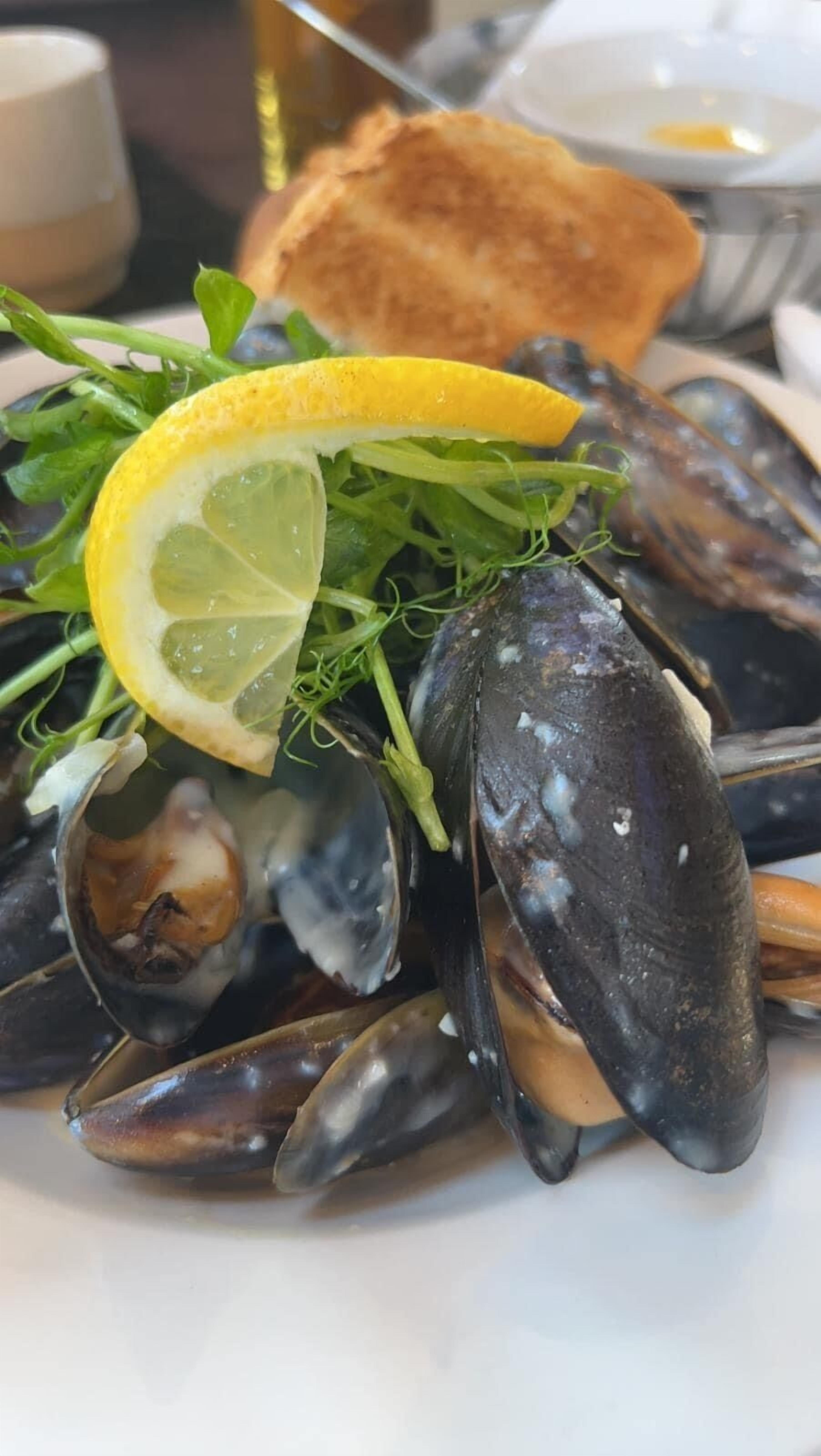 Une assiette de moules garnie d’une tranche de citron et de quelques feuilles de salade, accompagnée d’une tranche de pain grillé.