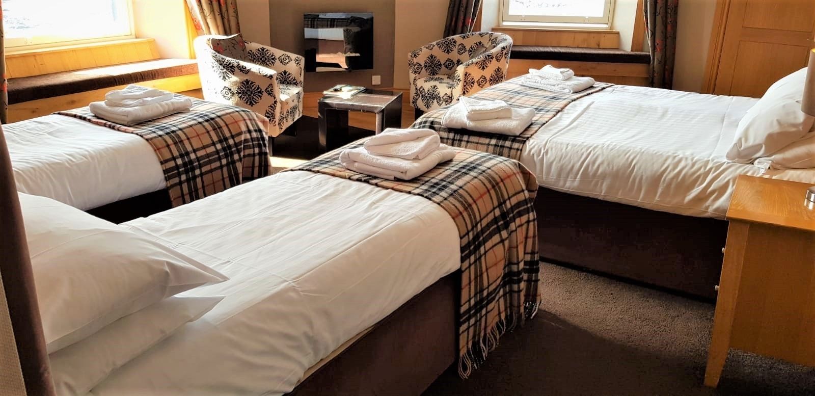 Een hotelkamer met twee bedden, elk met een ruitendekentje en witte lakens, een tafeltje met een lamp, en een stoel met een bloemenprint.