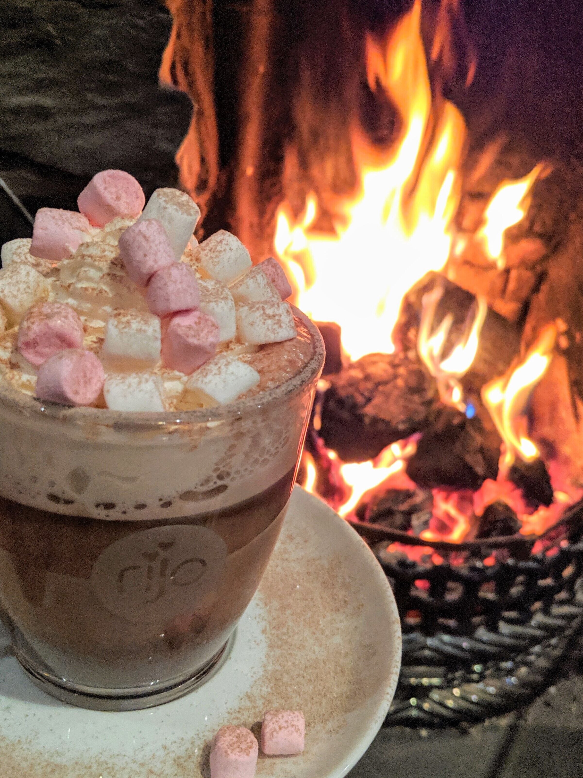 A hot beverage with marshmallows and a frothy top, served in a glass with a coaster, set against a backdrop of a roaring fire.