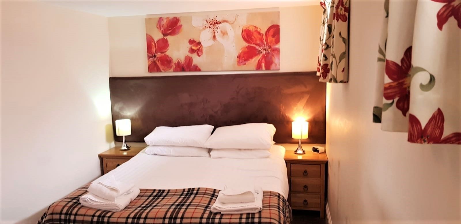 Una cama bien hecha con sábanas blancas, una manta escocesa y toallas dobladas al lado. Hay dos lámparas de noche y un cuadro de flores colgado en la pared sobre la cama.
