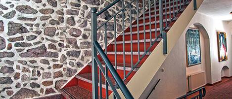 Una escalera con paredes de piedra y barandilla de metal, que conduce a una habitación con alfombra estampada y un cuadro en la pared.