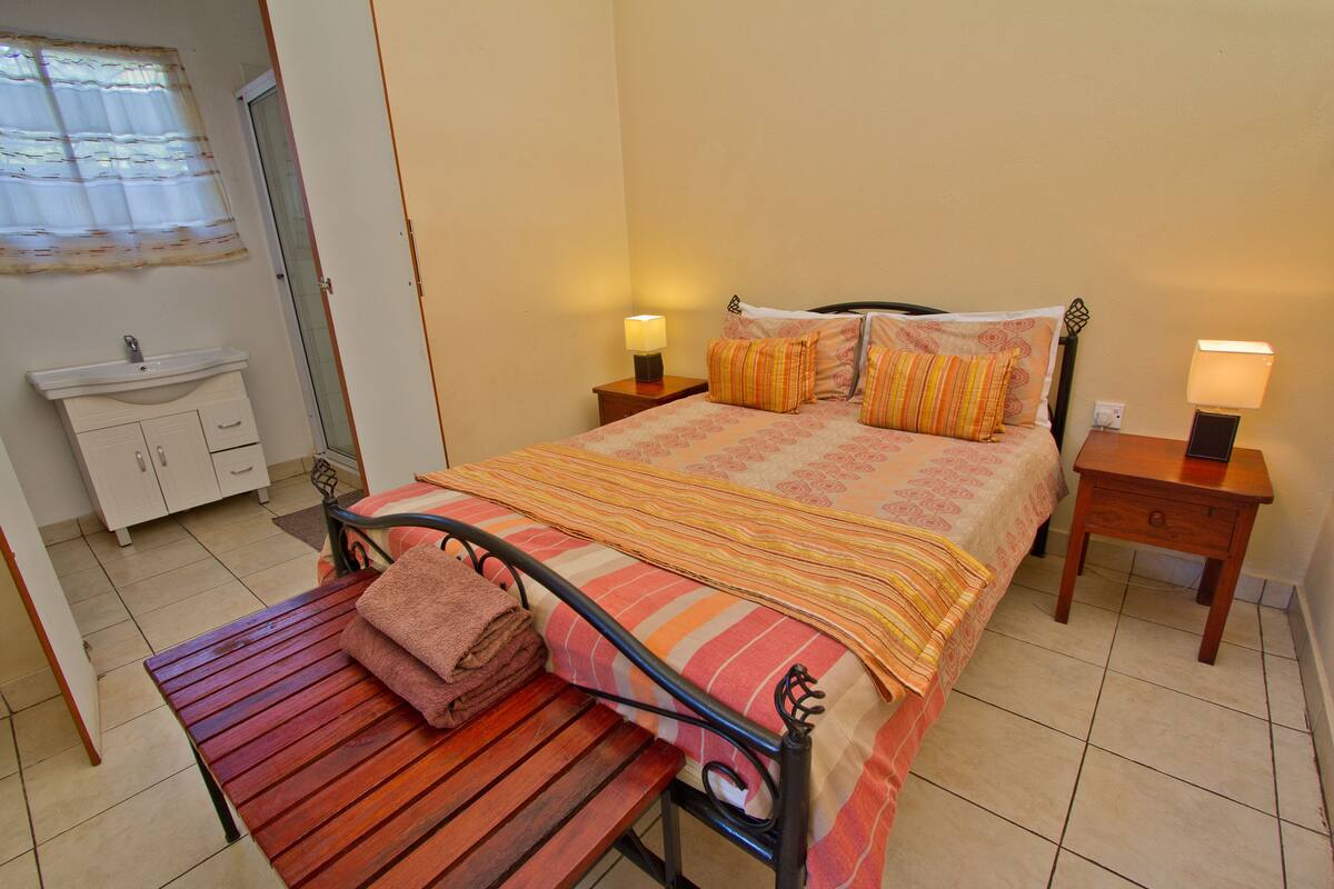 A bedroom with a bed, bedside tables, a nightstand, a bench, a mirror, and a bathroom with a sink and cabinets.