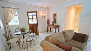 Uma sala de estar com sofá, mesa de jantar com cadeiras, porta de madeira e uma pequena mesa com uma planta.