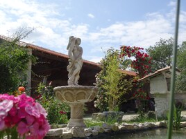 Um jardim com uma fonte de pedra, uma estátua e um edifício com telhado de azulejos.