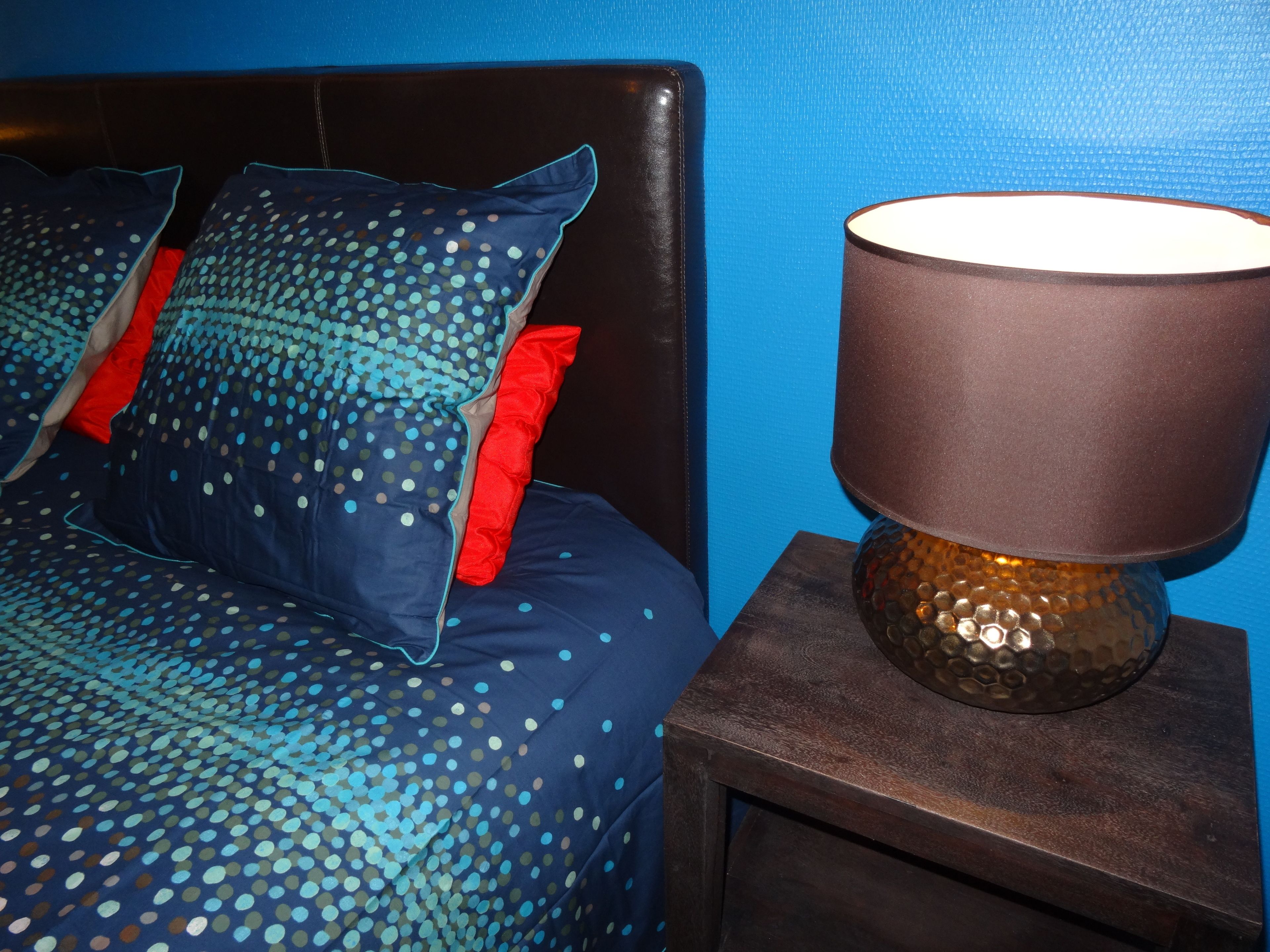 A bed with a polka dot cover and red pillows, a dark wooden headboard, and a lamp on a wooden nightstand.