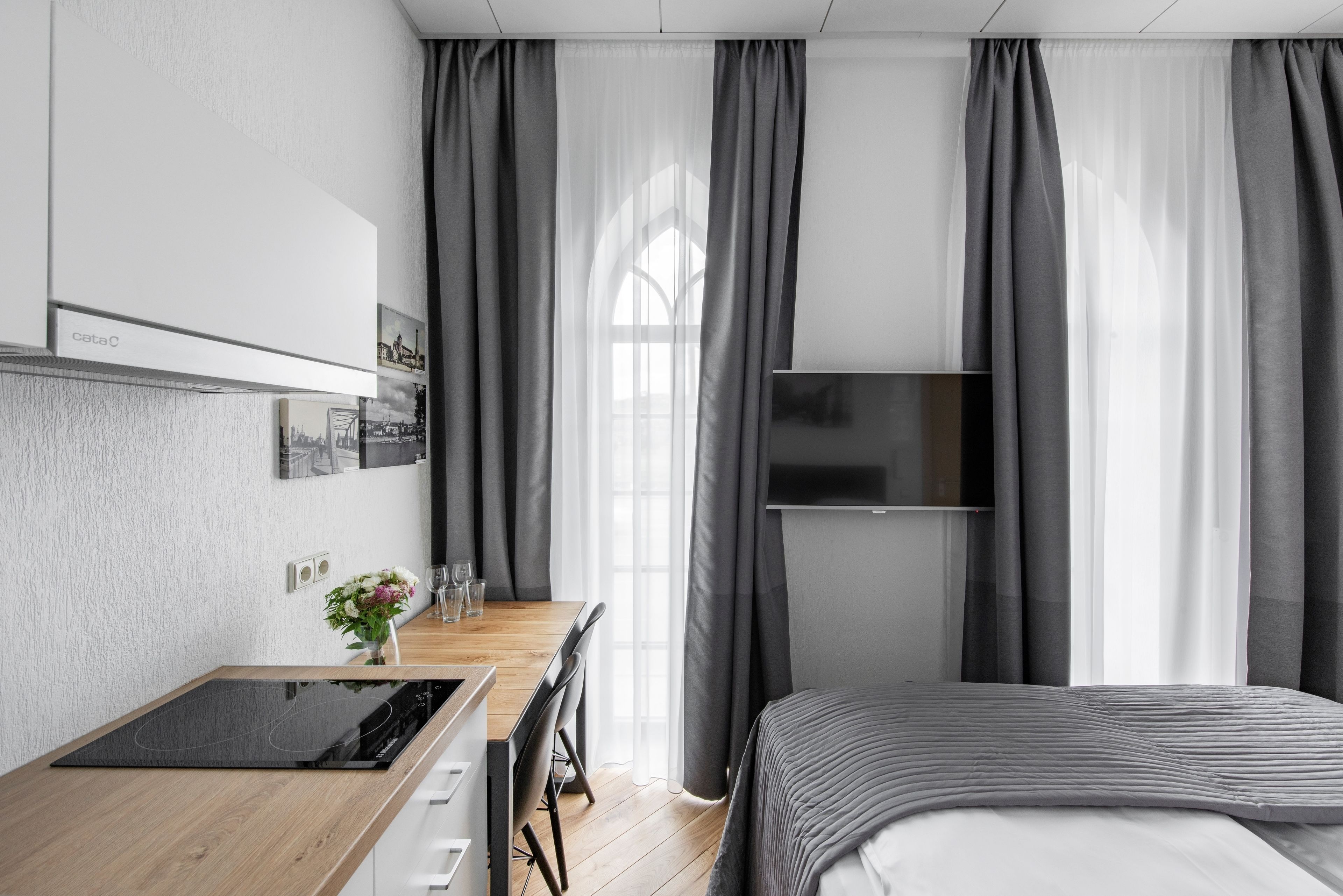 Ein modernes Hotelzimmer mit einem Holztisch, einem Flachbildfernseher, einem Bett mit grauer Tagesdecke und einem großen Fenster mit transparenten Vorhängen.