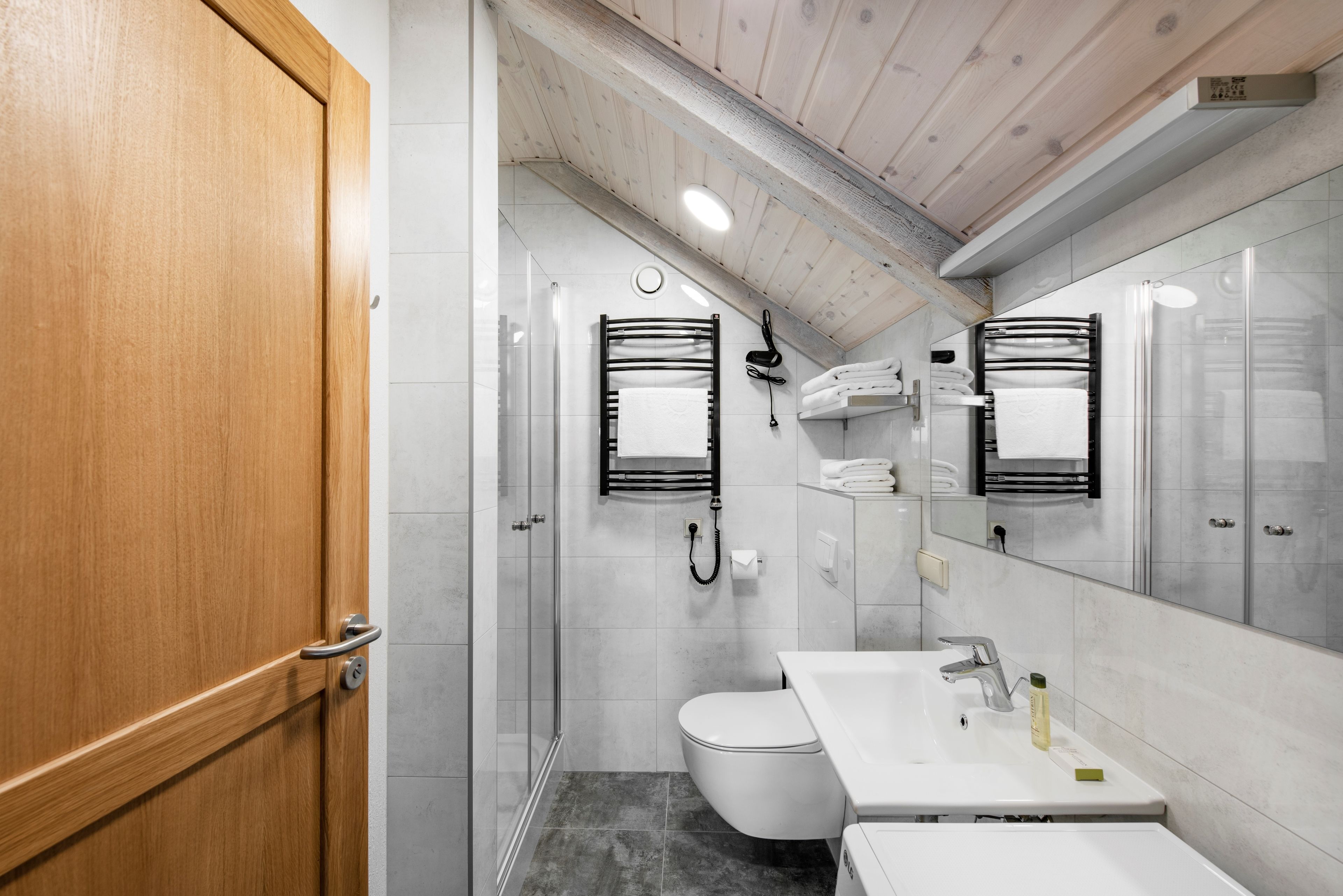 Un baño moderno con puerta de madera, cabina de ducha de vidrio, inodoro blanco, lavamanos con espejo y toalleros.