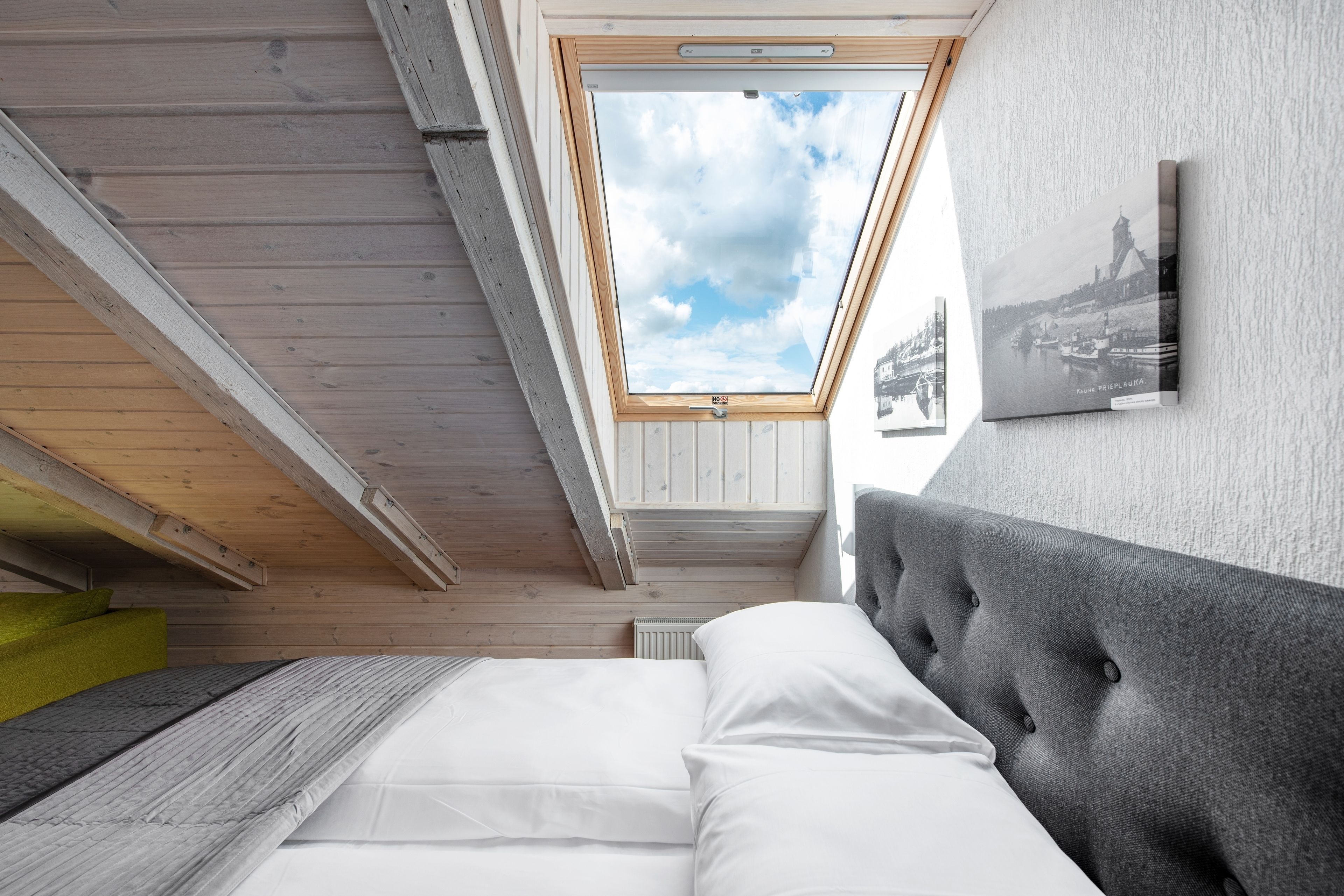 Una habitación con tragaluz, una cama con ropa de cama blanca, un cabecero tapizado en gris y un cuadro enmarcado en la pared.