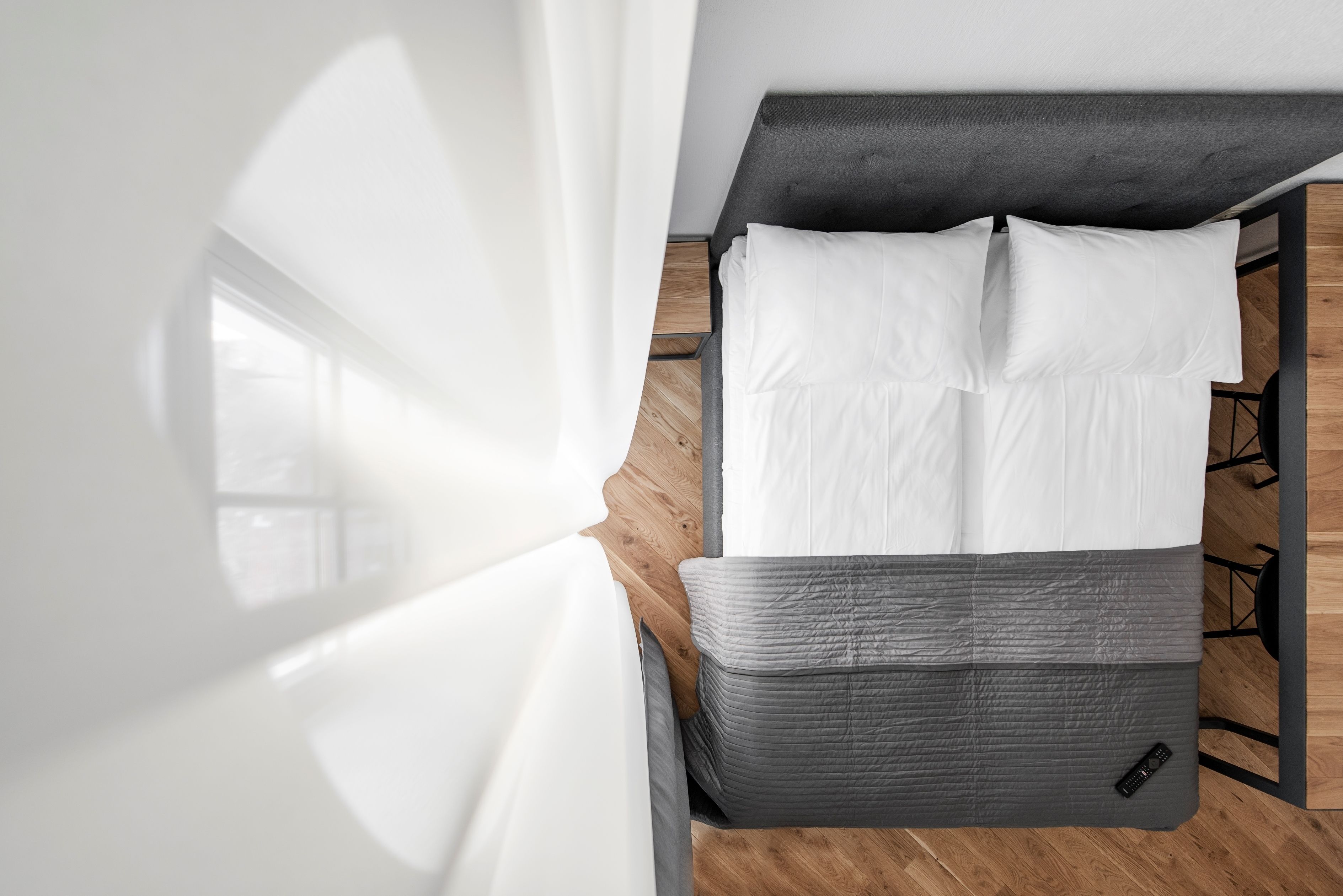 Cama bem arrumada com roupa de cama branca e cinza, piso de madeira e cabeceira moderna.