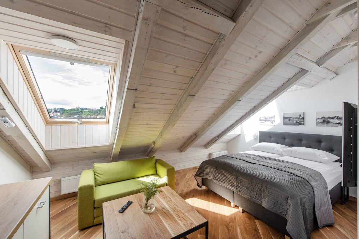 A room with a sloped ceiling, a bed, a green sofa, a wooden table, and a window with a view of greenery.