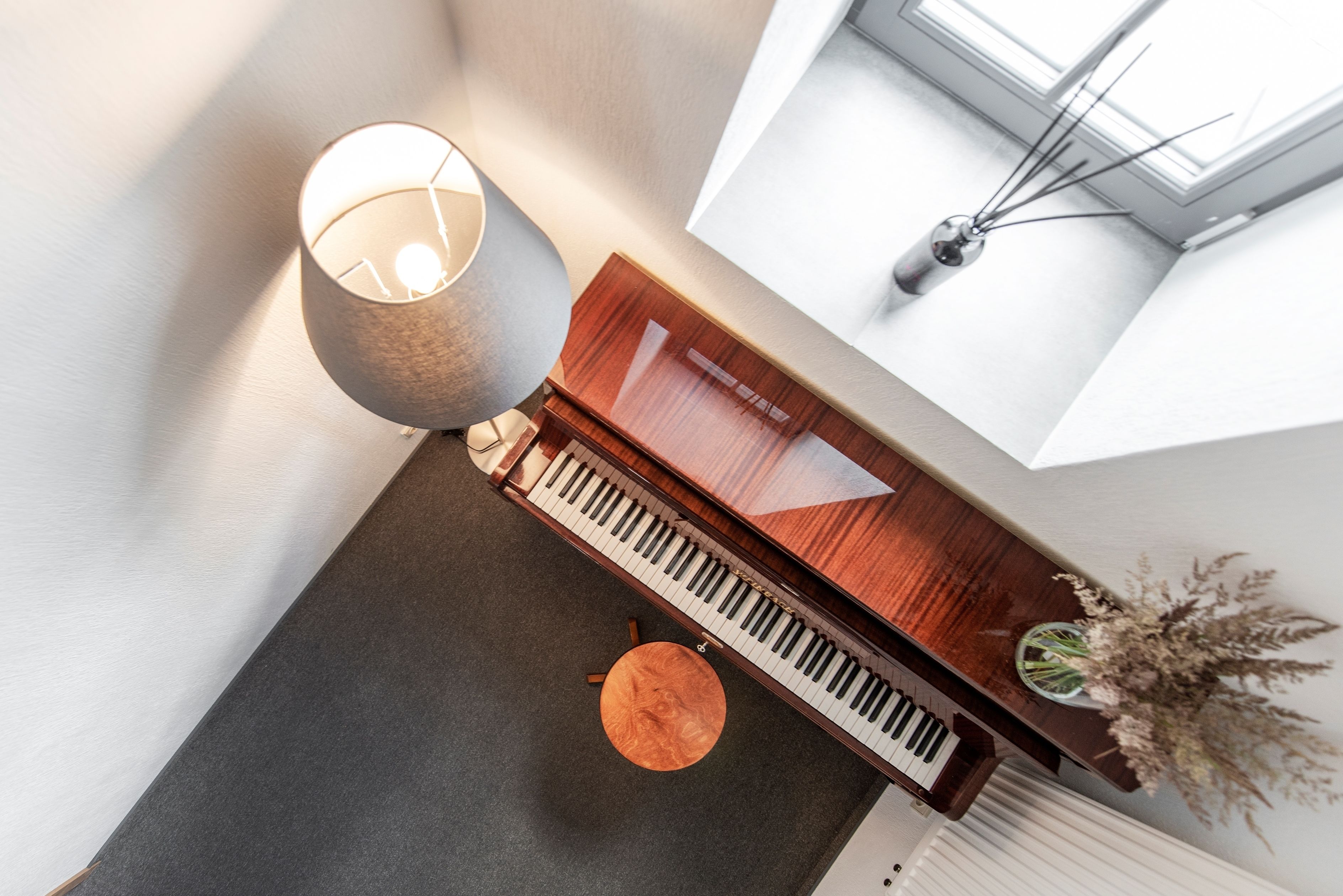 A piano, a lamp, and a vase with dried flowers on a table in a room with a window and a plant.