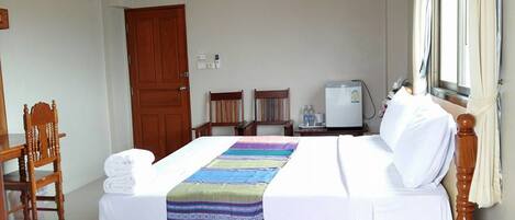 Quarto de hotel com cama de madeira, roupa de cama branca, manta colorida, escrivaninha com cadeira, mesa pequena, frigorífico e janela com cortinas.