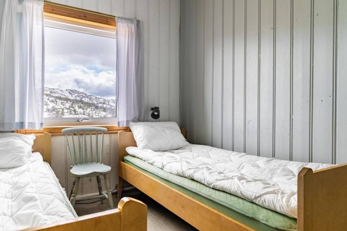 A small, single-room accommodation with two wooden bunk beds, a chair, a window with a view of a snowy landscape, and a wall-mounted clock.