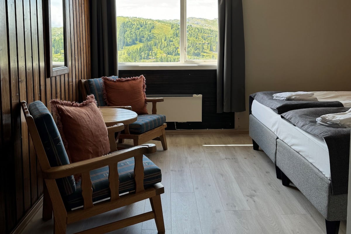A room with a wooden floor, a bed, a window with a view of green hills, a ceiling fan, and a radiator.