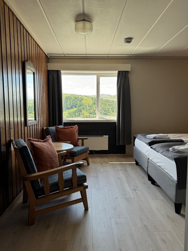 A room with a wooden floor, a bed, a window with a view of green hills, a ceiling fan, and a radiator.
