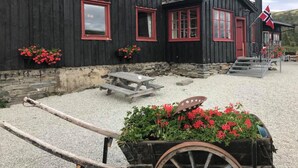 A black wooden building with red trim and windows, a flag, and a cart with flowers in front.