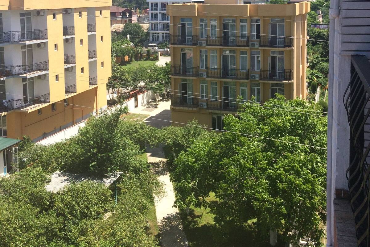 A multi-story yellow building with balconies, surrounded by greenery and a hillside in the background.