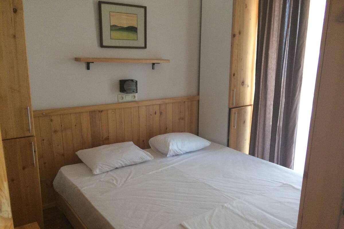 A single bed with white linens in a room with wooden paneling and a shelf above it.