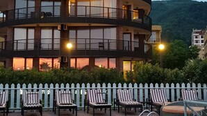 A multi-story building with balconies, a pool area with lounge chairs, and a white picket fence.