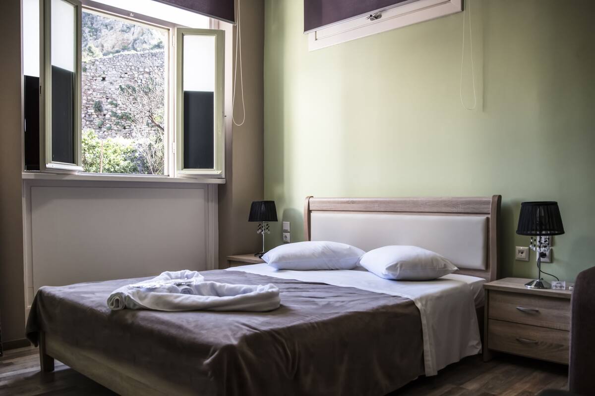 A neatly made bed with pillows, a bedside lamp, and a nightstand in a room with a window overlooking greenery.
