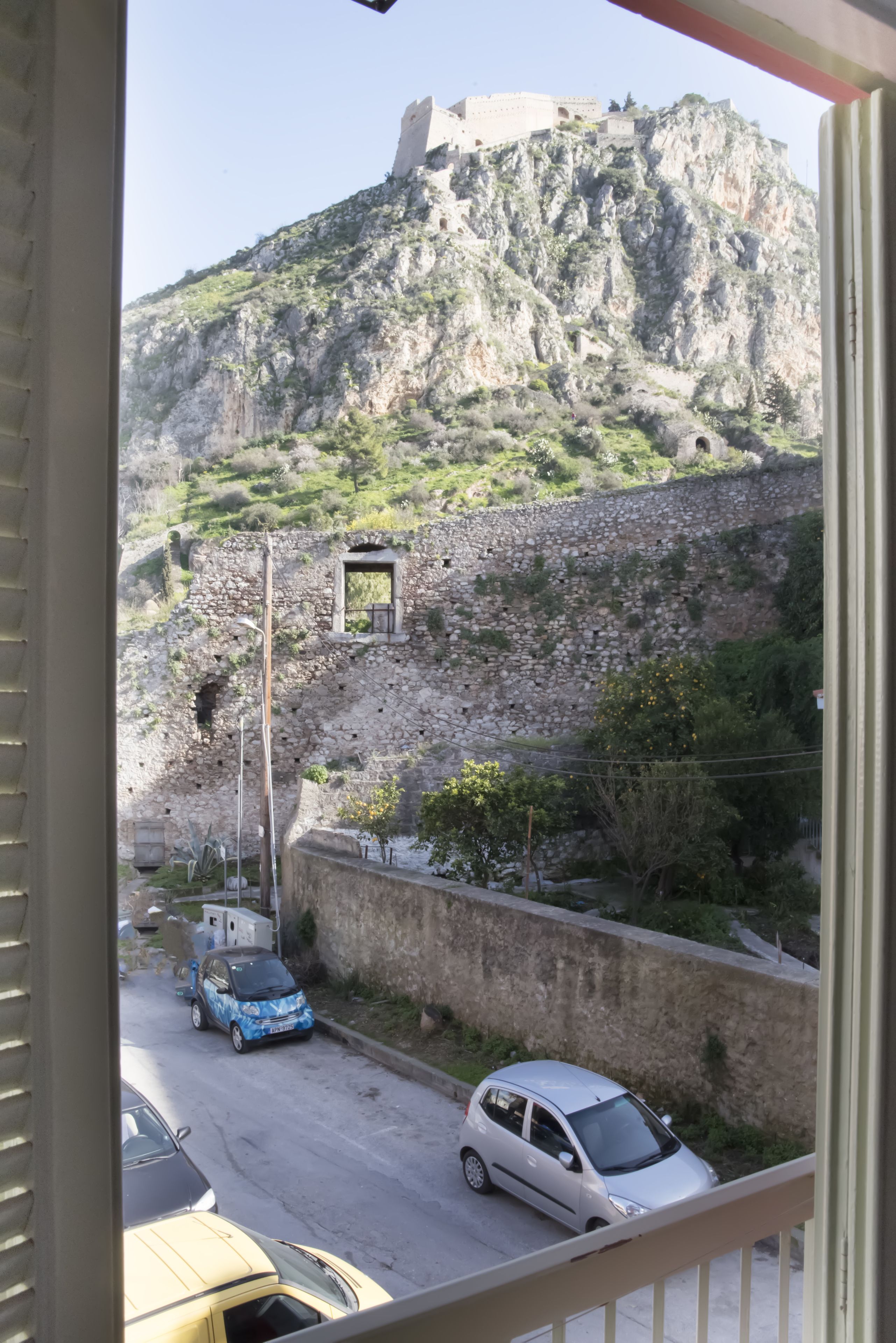 Vista de una zona montañosa con un muro de piedra y una pequeña ventana. Se ven coches aparcados a lo largo de una calle en primer plano.