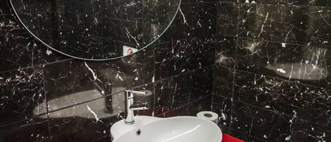 A bathroom with a white sink on a red countertop, a mirror above the sink, and black marble walls.