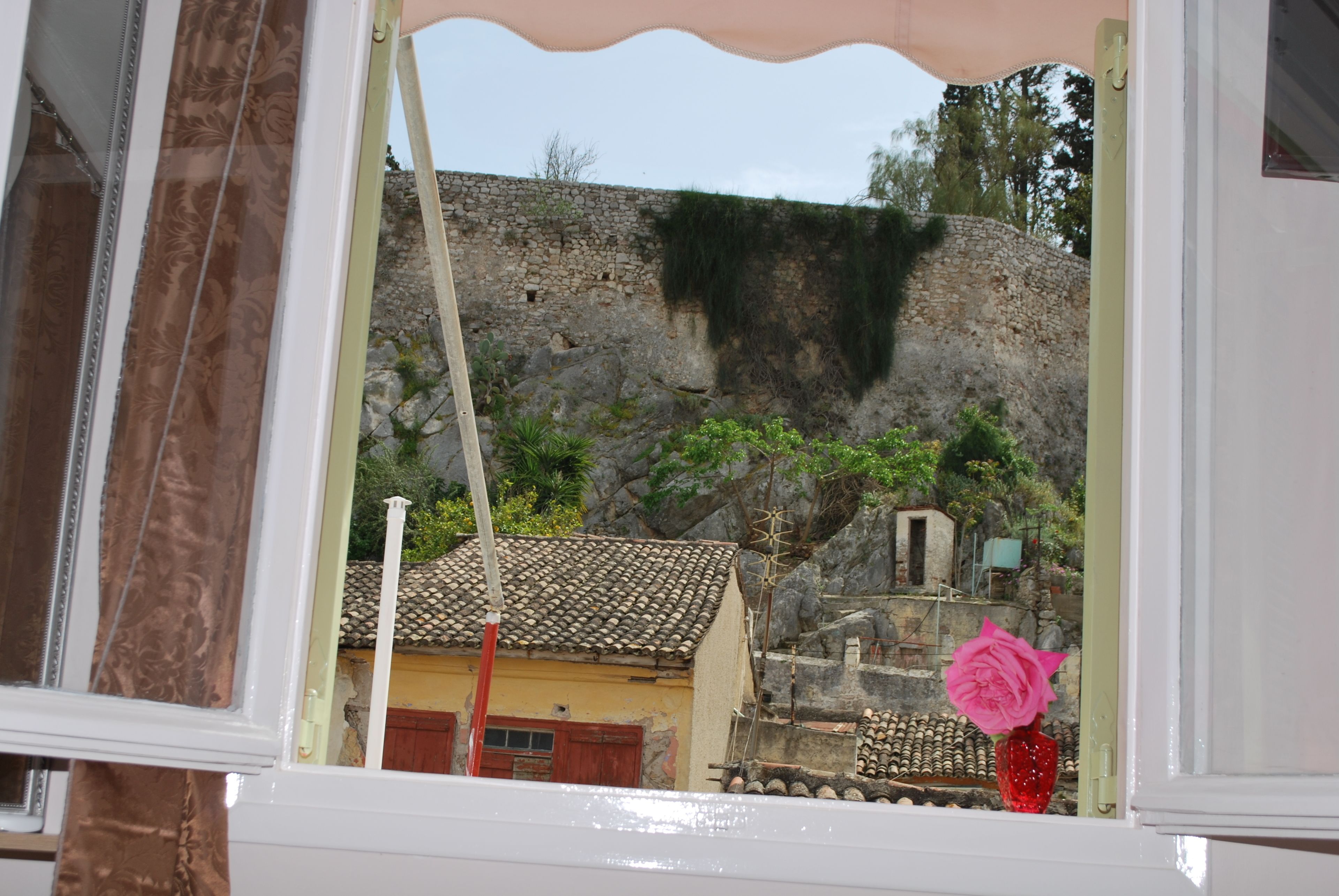 A view through a window of a building with a tiled roof, a stone wall, and a pink rose in a red vase.