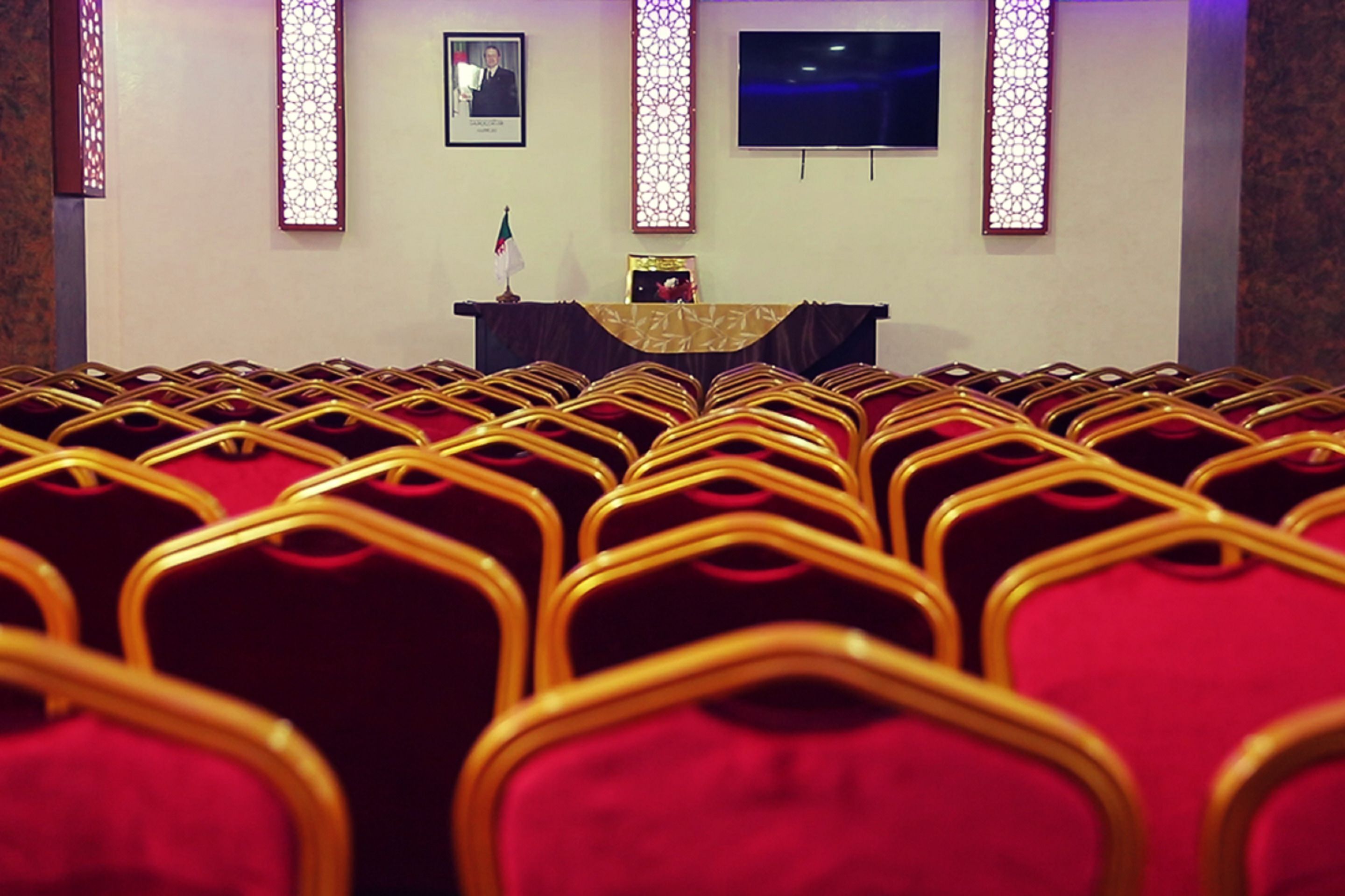 Rows of red chairs with golden frames in a room with a stage, a framed portrait, and decorative light panels.
