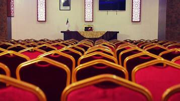Rows of red chairs with golden frames in a room with a stage, a framed portrait, and decorative light panels.