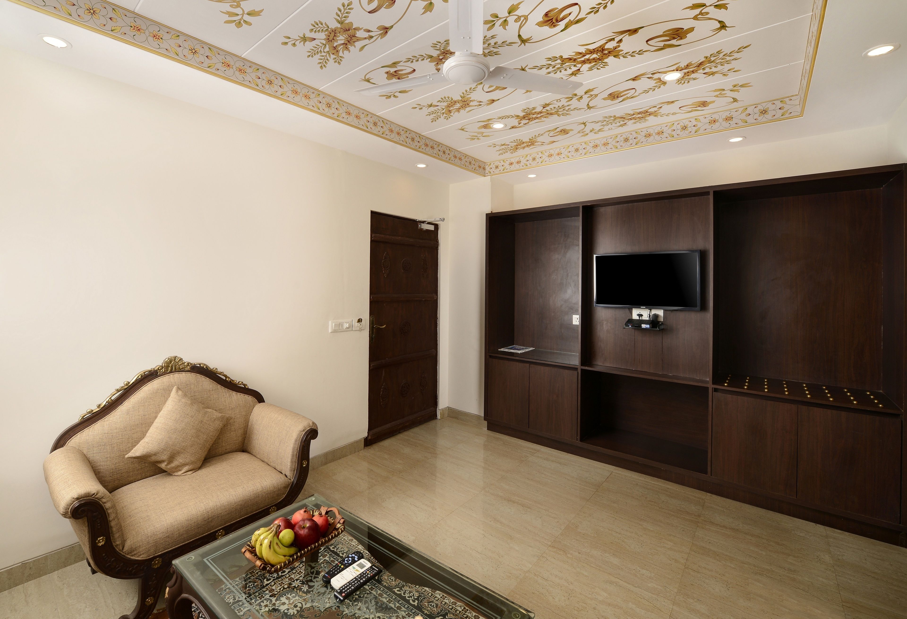 A living room with a sofa, a coffee table with fruits and a remote, a TV on a wooden cabinet, and a decorative ceiling.