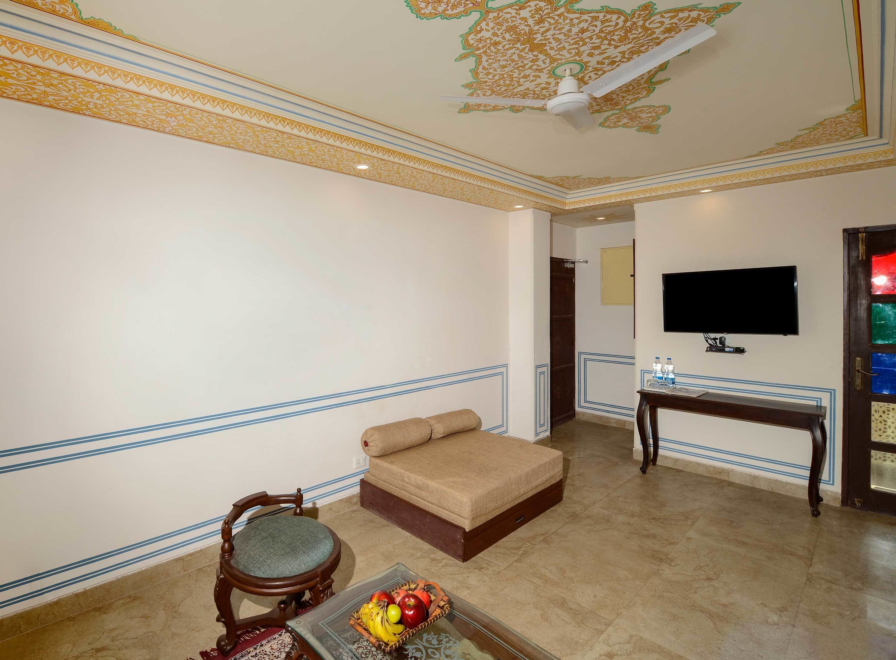A hotel room with a sofa, a wooden chair, a small table with fruit, a television, and a decorative ceiling.