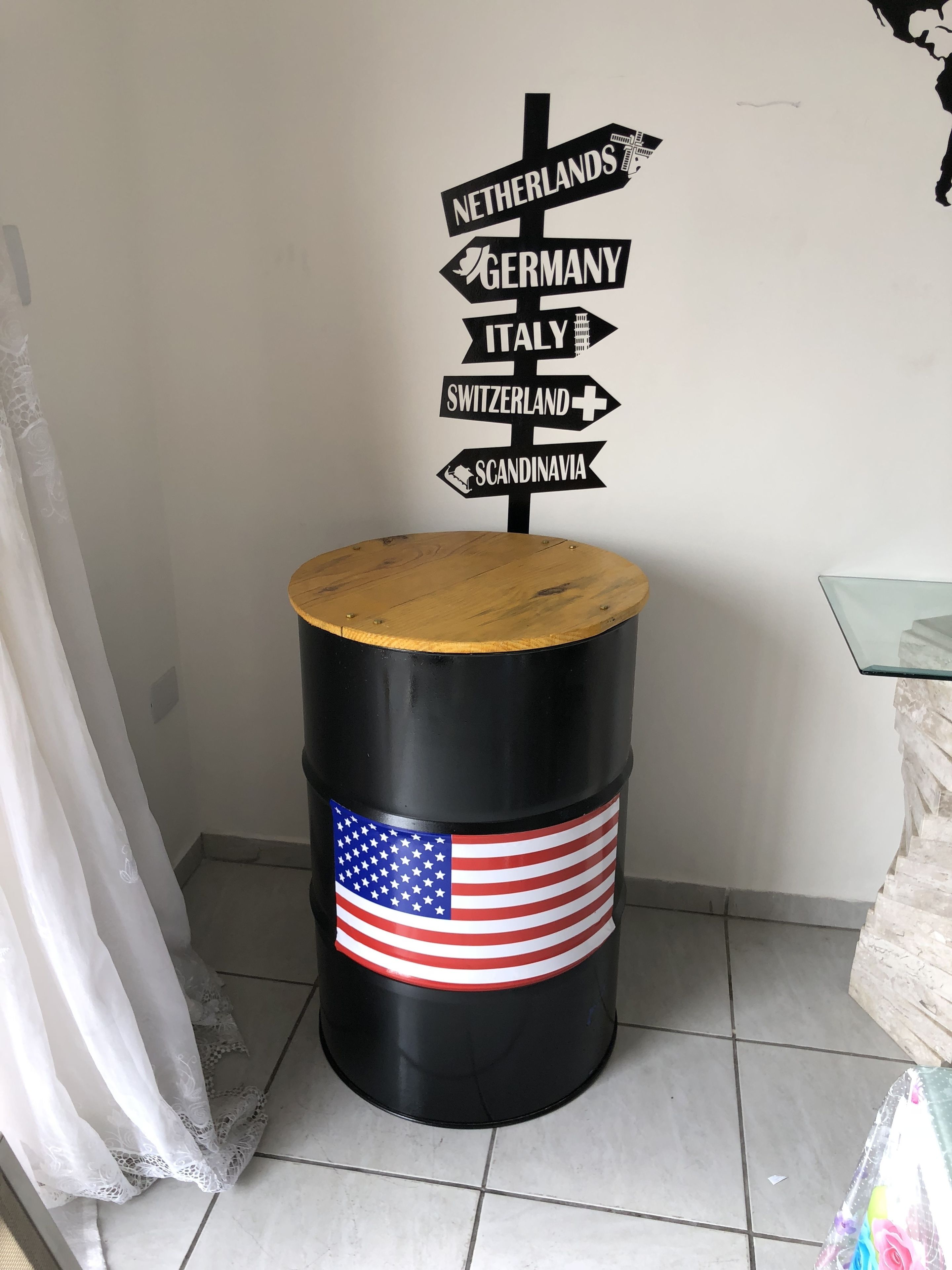 Um barril preto com tampa de madeira e uma bandeira americana pintada, posicionado contra uma parede branca com placas de sinalização indicando vários países.