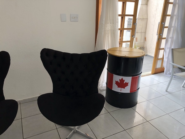 Una habitación con una silla tapizada de negro, un barril negro con una hoja de arce roja y una puerta blanca con un ventanal.