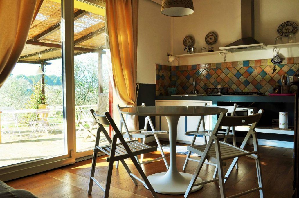Un espace repas avec une table ronde et des chaises, une cuisine agrémentée d’un crédence colorée, et une vue sur l’extérieur grâce à de grandes fenêtres.