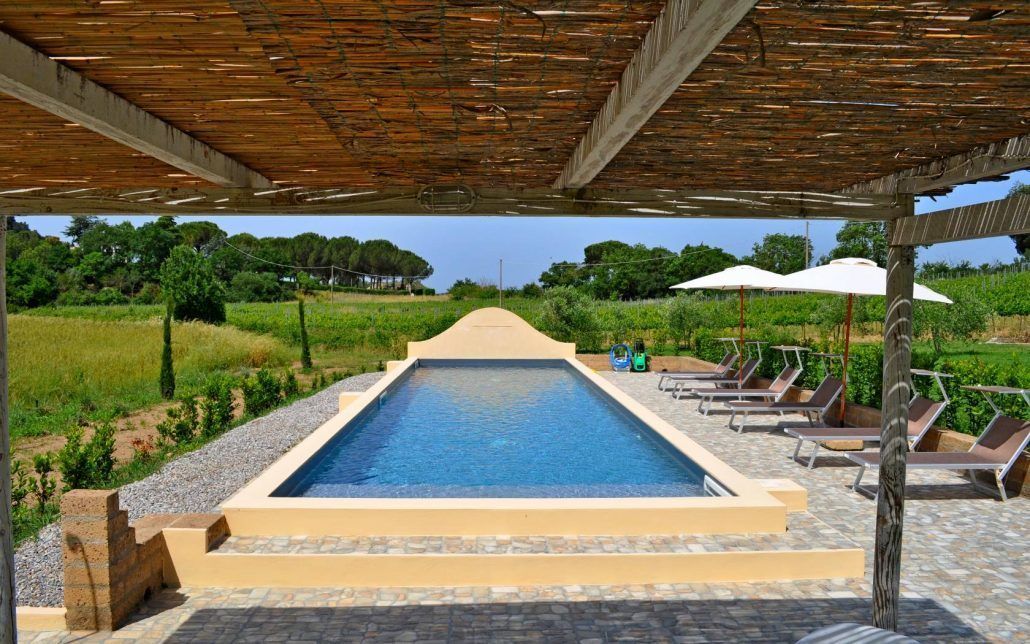 Una piscina rettangolare con bordo piastrellato, circondata da lettini e ombrelloni, sotto un pergolato in legno.