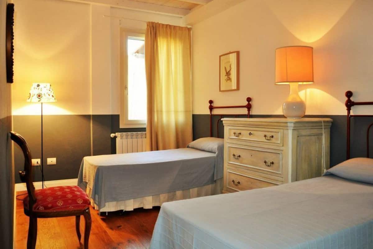 A room with two single beds, a wooden dresser, a chair, a lamp, a window with curtains, and a radiator.