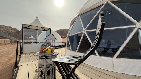A desert camp with a large dome tent, a small pavilion, and a wooden deck with a table and fruits.