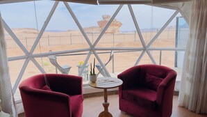 A room with a large glass window offering a view of a desert landscape. Inside, there are two red velvet chairs and a small round table with a nameplate. - Mazayen Rum Camp (Wadi Rum)