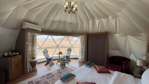 A tent-style room with a bed, two chairs, a small table, and a view of a desert landscape. - Mazayen Rum Camp (Wadi Rum)