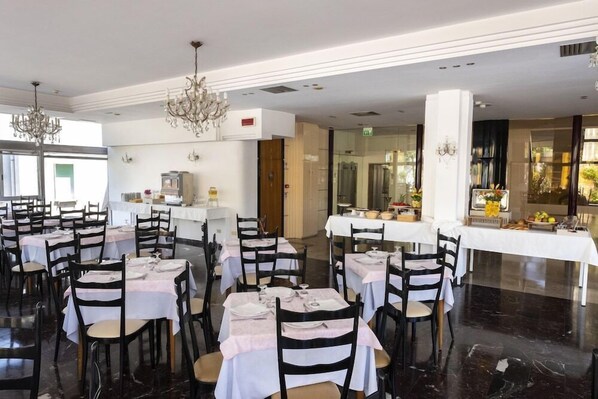 Uma sala de jantar ampla com várias mesas e cadeiras, um lustre e um buffet.