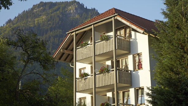 Un edificio a più piani con balconi, circondato da alberi e montagne.
