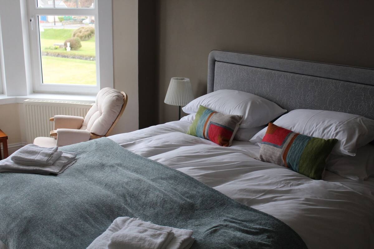 A neatly made bed with white linens and colorful pillows, a grey headboard, and a chair with a white cushion.