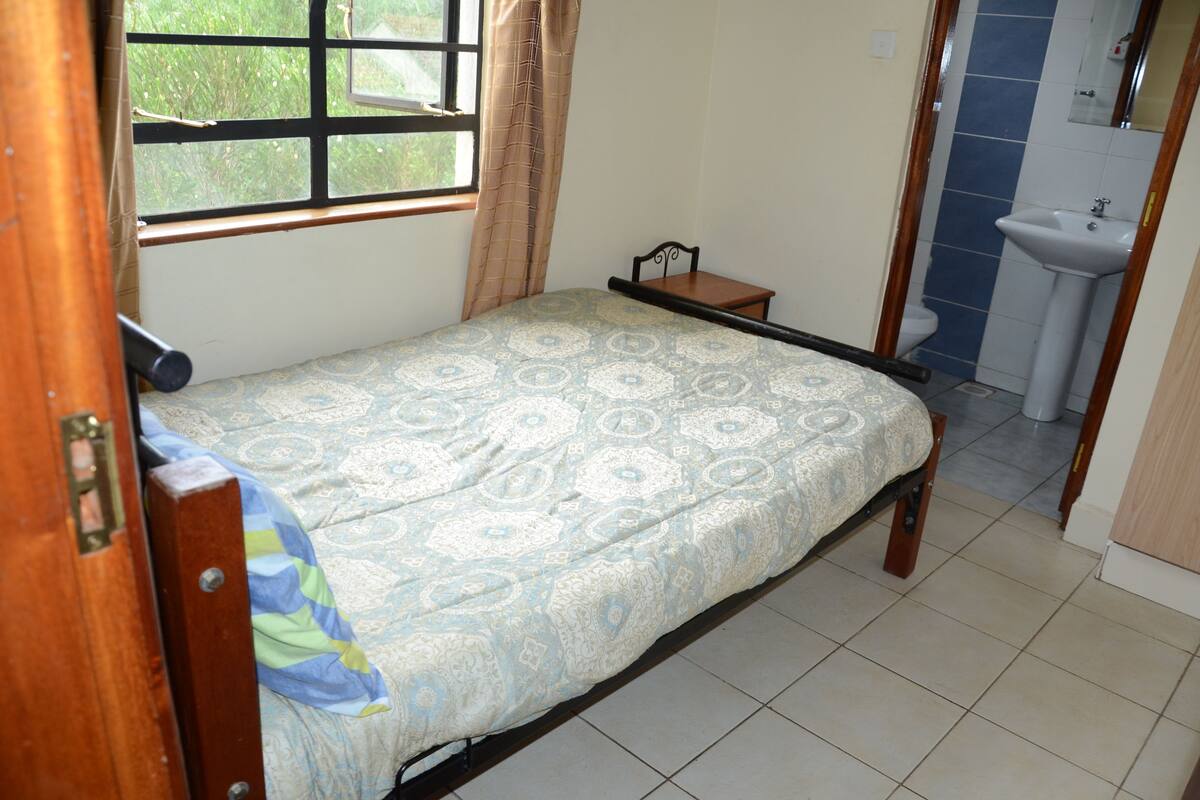 A small, single-bed room with a patterned bedspread, a window with curtains, a small bedside table, and a bathroom with a sink and toilet.