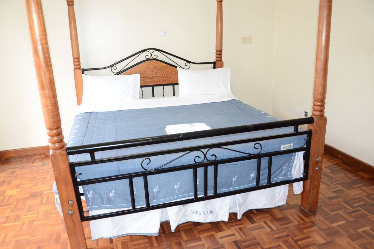 A four-poster bed with a wooden headboard, white bedding, and a blue bedspread with a "She's Club" logo.