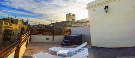 A rooftop terrace with a white lounge chair, a small table, and a trash bin. The terrace has a view of the cityscape and a historic building in the background.
