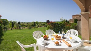 A round table with white chairs set outdoors, surrounded by a lush garden and a clear blue sky. - Vacancéole - Port Minervois (Homps)