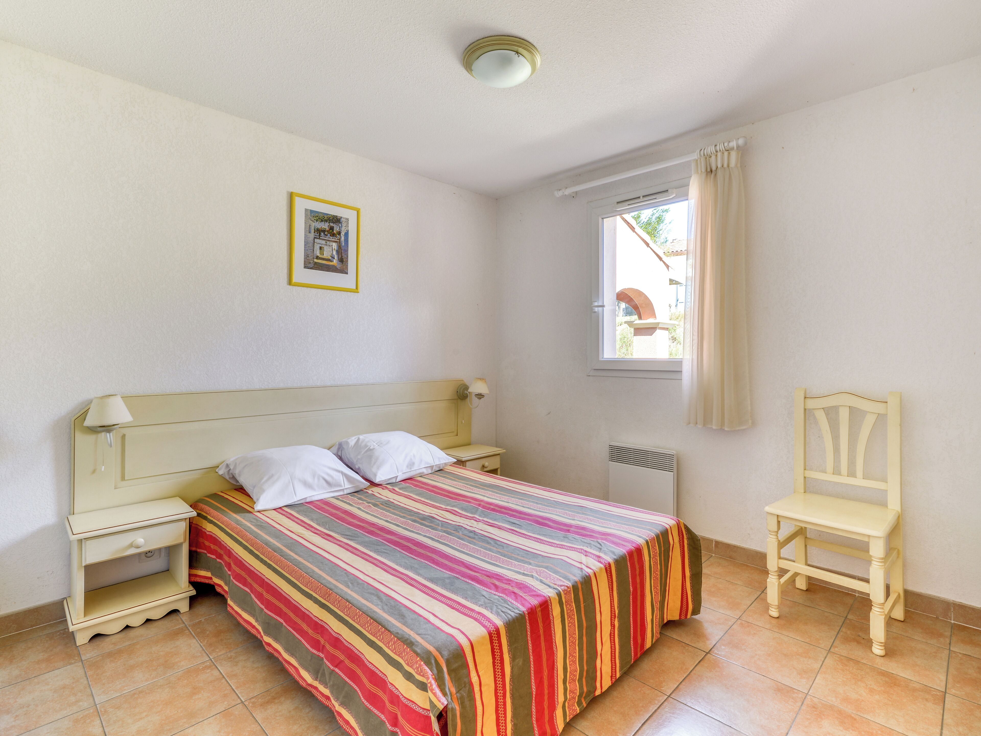 A bedroom with a bed, bedside table, chair, window, and a picture on the wall.