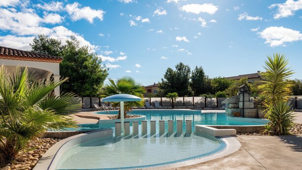 A swimming pool with a curved edge, surrounded by palm trees and a fence. - Vacancéole - Port Minervois (Homps)